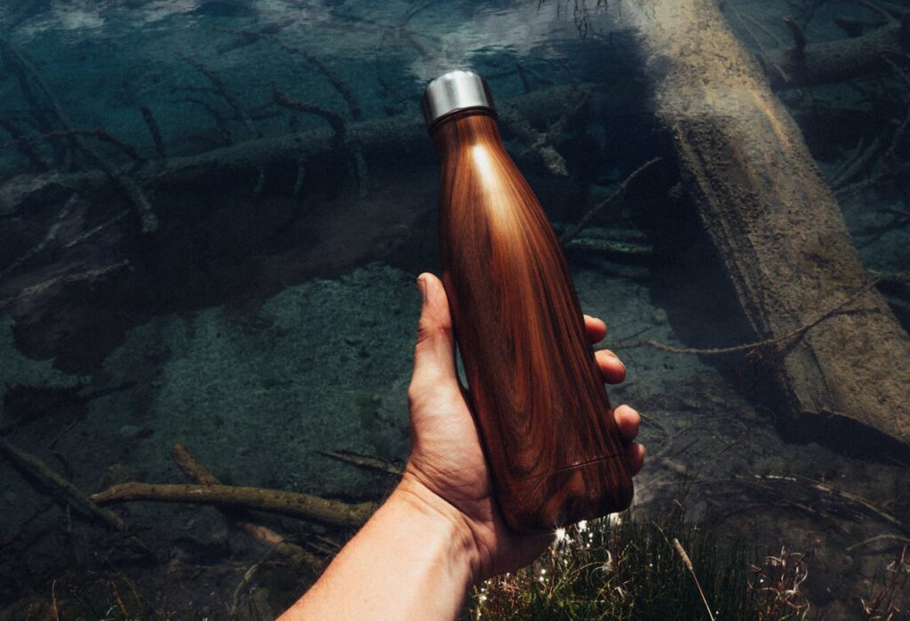 A hand holding a stainless steel wood-grain water bottle over a clear stream.