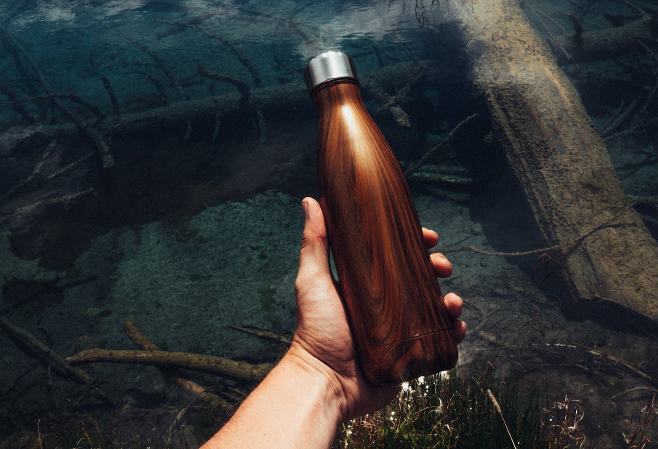 A hand holding a stainless steel wood-grain water bottle over a clear stream.